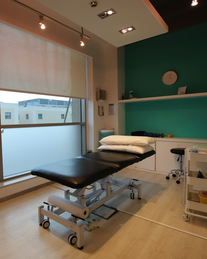 Behandlungsraum mit schwarzer Therapieliege, zwei Kissen, Rollhocker und einer salbeigrünen Wand mit Uhr